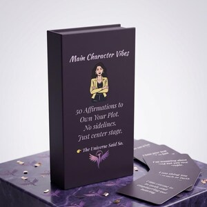 May include: A dark purple rectangular box featuring the text Main Character Vibes, an illustration of a person with crossed arms, and the text 50 Affirmations to Own Your Plot. No sidelines. Just center stage. The Universe Said So. with a phoenix logo.