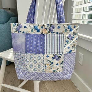 May include: A patchwork tote bag with a floral and geometric pattern in shades of blue, purple, and cream. The bag features a wooden button closure and matching patterned handles. The bag is sitting on a white chair.