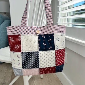 May include: A patchwork tote bag with a patriotic theme. The bag features a variety of fabric squares in red, white, and blue patterns, including stars, floral designs, and gingham. The handles and top border are red and blue gingham. A wooden button secures the top.