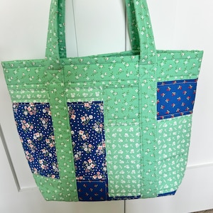 May include: A quilted tote bag with a patchwork design. The bag features a combination of green, navy blue, and white floral patterns. The bag has two sturdy handles and is suitable for carrying various items.