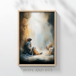 Puede incluir: Una impresión en acuarela enmarcada representa una figura con una túnica y un tocado, sentada en un entorno similar a una cueva, mirando hacia arriba hacia una luz brillante. La obra de arte está en tonos apagados de beige, azul y dorado. El texto "HOPE AND HUE" está impreso en la parte inferior.