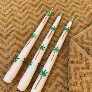 Puede incluir: Tres velas blancas de tipo taper con diseños de palmeras verdes. Las velas están sobre una superficie marrón texturizada.