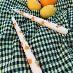 Pode incluir: Duas velas brancas com desenhos de frutas laranja sobre uma toalha de mesa quadriculada verde e preta. As velas são longas e finas, e os desenhos de frutas são simples e estilizados.