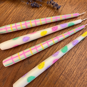 Może przedstawiać: Cztery długie, białe świece stożkowe z pastelowymi wzorami w kratkę w kolorze różowym, zielonym i żółtym oraz kolorowymi kropkami. Świece są ułożone na drewnianej powierzchni.