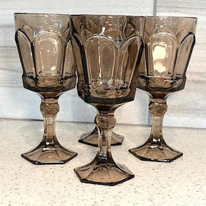 May include: Four vintage brown-tinted glass goblets. Each goblet features a faceted bowl, a decorative stem, and a hexagonal base. The glasses are arranged on a light-colored surface, showcasing their elegant design and color.