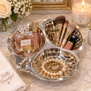 May include: A silver-toned, three-sectioned shell-shaped vanity tray holding a Miss Dior perfume bottle, a lip gloss, makeup brushes, an eyelash curler, a lipstick, and a pearl bracelet with a gold ring. The tray has a metallic finish.