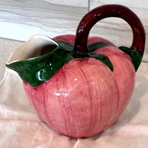 May include: A ceramic pitcher with a bulbous, pink body and a dark brown handle. The pitcher has green leaf accents and a white interior. The design resembles a stylized fruit, possibly a tomato or apple. The pitcher is sitting on a white cloth.