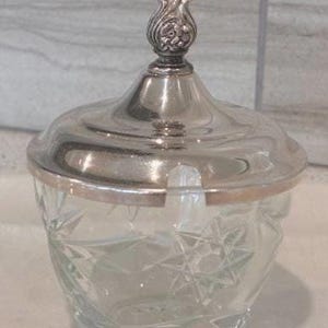 May include: A clear glass sugar bowl with a silver-colored lid and finial. The bowl has a cut-glass design, and the lid has a decorative floral design on top. The sugar bowl is a classic piece for serving sugar or other condiments.
