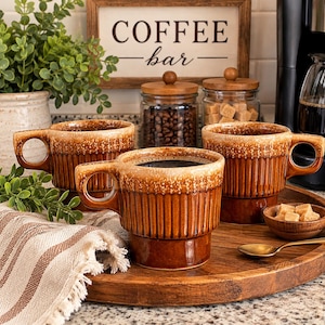 May include: Three brown ceramic mugs with a vertical ribbed texture and a cream-colored drip glaze around the rim. Each mug features a wide handle and a stacked base design, arranged on a round wooden tray with a small wooden bowl of sugar cubes.