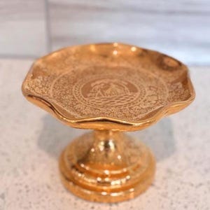May include: A gold-colored, pedestal cake stand with a scalloped edge. The stand features an ornate, embossed design on the plate and base. The plate has a central medallion with a landscape scene.