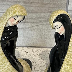 May include: Two ceramic figurines of women in black robes and yellow textured hoods. The faces are painted with delicate features, including closed eyes and rosy cheeks. The figures are in a seated position.