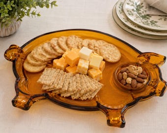 May include: An amber-colored glass serving tray shaped like a pig, featuring a built-in circular bowl on the right side. The tray is filled with round crackers, square crackers, cubes of orange and white cheese, and a small bowl of mixed nuts.