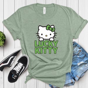 Peut inclure: T-shirt vert sauge avec un chat de dessin anim&eacute; blanc, un n&oelig;ud vert et les mots "LUCKY KITTY" en vert. L'image comprend des baskets noires et blanches et un short en jean.