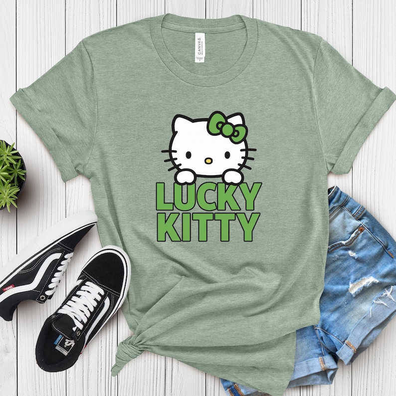 Peut inclure: T-shirt vert sauge avec un chat de dessin anim&eacute; blanc, un n&oelig;ud vert et les mots "LUCKY KITTY" en vert. L'image comprend des baskets noires et blanches et un short en jean.