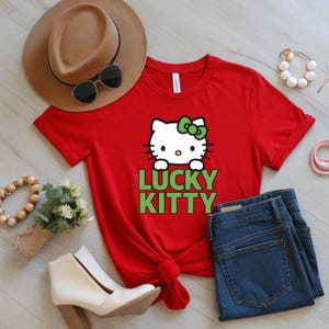 Peut inclure: T-shirt rouge avec un chat de dessin anim&eacute; blanc, un n&oelig;ud vert et le texte "LUCKY KITTY" en vert. L'image comprend un jean bleu, un chapeau marron, des lunettes de soleil et des bottines blanches.