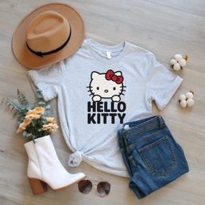 Peut inclure: Un t-shirt gris clair avec un motif Hello Kitty, un n&oelig;ud rouge et le texte "HELLO KITTY" en noir. Le t-shirt est associ&eacute; &agrave; un jean bleu, un chapeau beige, des bottes blanches et des lunettes de soleil.