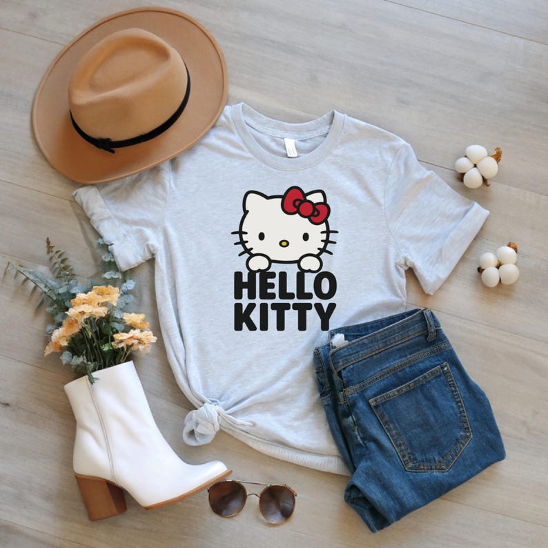 Peut inclure: Un t-shirt gris clair avec un motif Hello Kitty, un n&oelig;ud rouge et le texte "HELLO KITTY" en noir. Le t-shirt est associ&eacute; &agrave; un jean bleu, un chapeau beige, des bottes blanches et des lunettes de soleil.