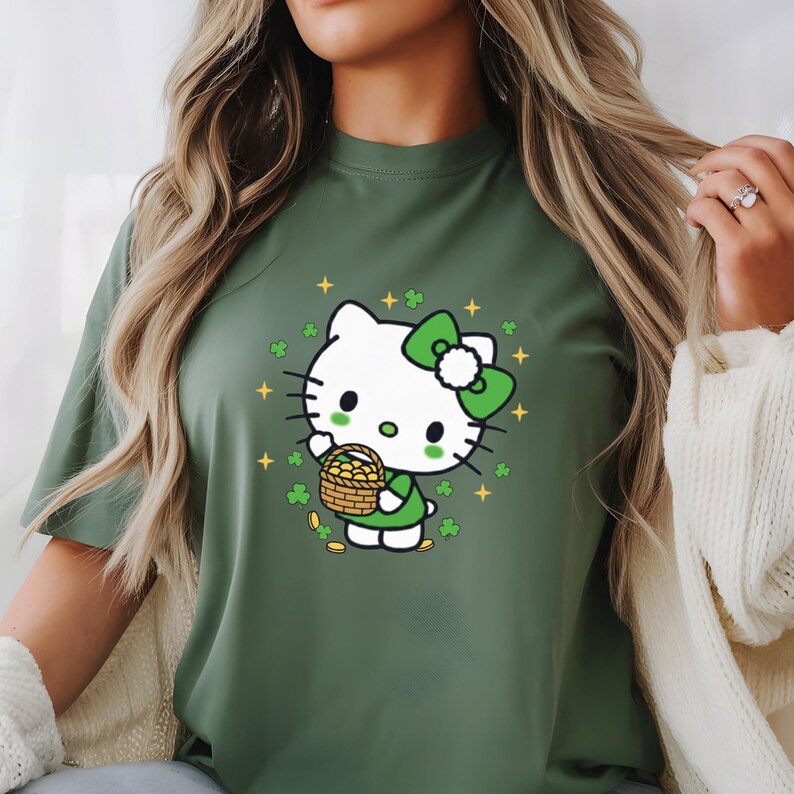 May include: Green t-shirt featuring a cartoon character with a white head, green bow, and green outfit, holding a basket of gold coins. The design includes shamrocks and gold stars, celebrating St. Patrick's Day.
