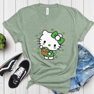 May include: A light green t-shirt featuring a cartoon character with a green bow, holding a basket of gold coins. The design includes green shamrocks and gold stars. The shirt is paired with denim shorts and black and white sneakers.