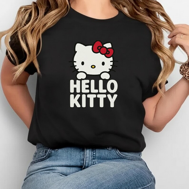 Peut inclure: T-shirt noir avec le personnage Hello Kitty. Le motif blanc pr&eacute;sente le visage de Hello Kitty avec un n&oelig;ud rouge et l'inscription "HELLO KITTY" en lettres blanches. Le t-shirt est de style col rond.