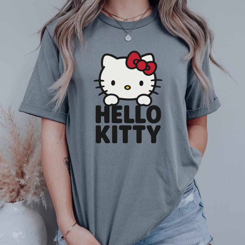 Peut inclure: Un t-shirt gris avec un motif Hello Kitty. Le motif pr&eacute;sente un visage de chat blanc avec un n&oelig;ud rouge et les mots "HELLO KITTY" en lettres noires.