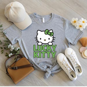 Peut inclure: T-shirt gris avec un motif Hello Kitty, un n&oelig;ud vert et l'inscription "LUCKY KITTY" en vert. L'image comprend une casquette de baseball beige, un sac &agrave; main marron et des baskets blanches.