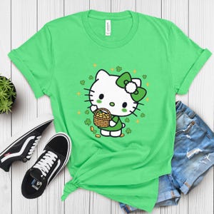 May include: A bright green t-shirt featuring a cartoon character with a green bow and dress, holding a basket of gold coins. The design includes shamrocks and gold sparkles. Black and white sneakers and denim shorts are also visible.