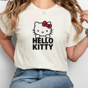 Peut inclure: T-shirt ivoire avec un motif Hello Kitty. Le motif pr&eacute;sente un visage de chat blanc avec un n&oelig;ud rouge et les mots "HELLO KITTY" en noir. Le t-shirt est en mati&egrave;re souple et est un v&ecirc;tement d&eacute;contract&eacute;.