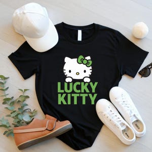 Peut inclure: T-shirt noir avec un motif Hello Kitty blanc, un n&oelig;ud vert et le texte "LUCKY KITTY" en vert. L'image comprend &eacute;galement une casquette de baseball blanche, des baskets blanches, des lunettes de soleil et une chaussure marron.