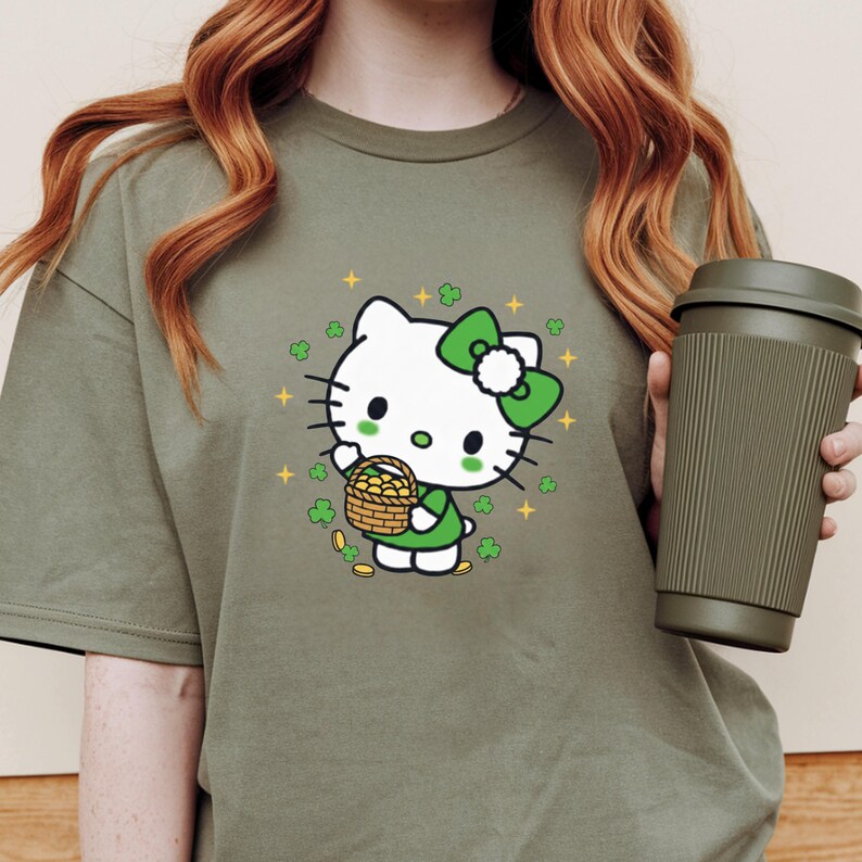 May include: Olive green t-shirt featuring a cartoon character with a white face, green bow, and green dress, holding a basket of gold coins. The design includes shamrocks and gold stars. A reusable coffee cup is held in the hand.