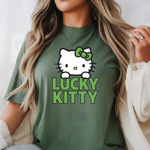 Peut inclure: T-shirt vert olive avec un chat de dessin anim&eacute; blanc orn&eacute; d'un n&oelig;ud vert et les mots "LUCKY KITTY" en vert. Le chat a des moustaches noires et un nez jaune. Le t-shirt est de couleur unie.