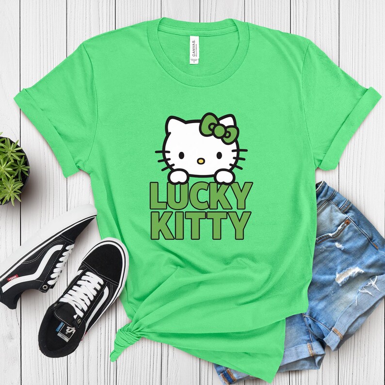 Peut inclure: Un t-shirt vert clair avec un motif de Hello Kitty avec un n&oelig;ud vert et le texte "LUCKY KITTY" en vert. Des baskets noires et blanches et un short en jean sont &eacute;galement visibles.