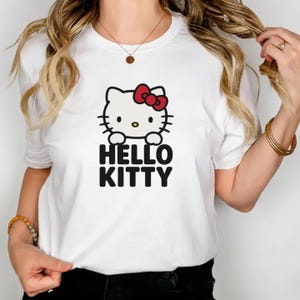 Peut inclure: T-shirt blanc avec un motif Hello Kitty. Le motif repr&eacute;sente un chat de dessin anim&eacute; avec un n&oelig;ud rouge et les mots "HELLO KITTY" en noir. Le t-shirt est de style col rond.