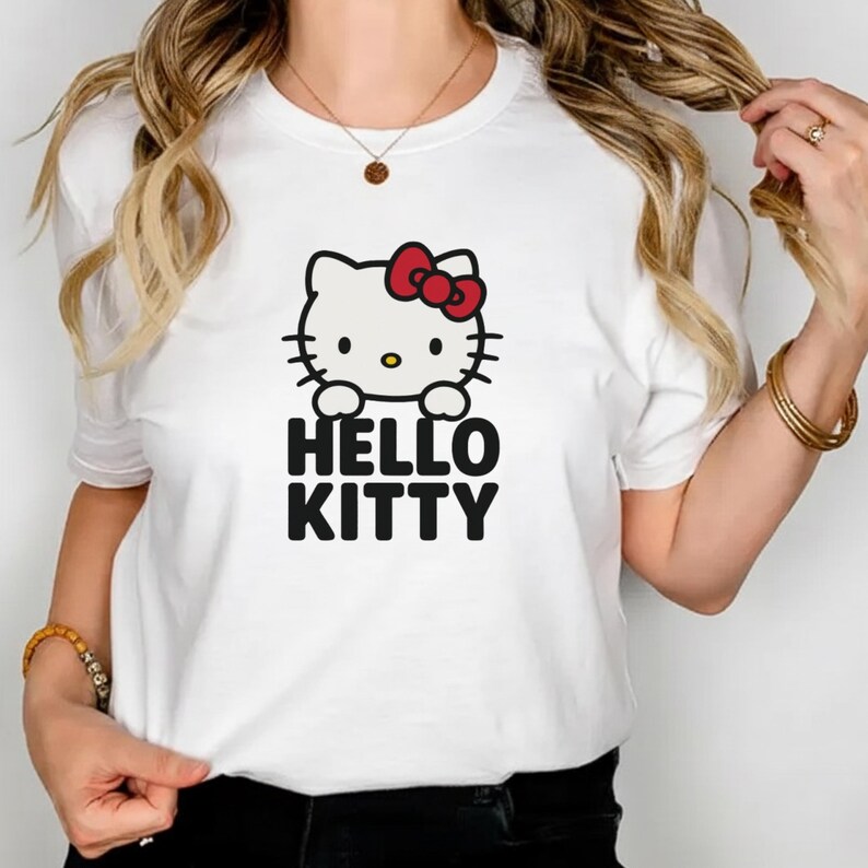 Peut inclure: T-shirt blanc avec un motif Hello Kitty. Le motif repr&eacute;sente un chat de dessin anim&eacute; avec un n&oelig;ud rouge et les mots "HELLO KITTY" en noir. Le t-shirt est de style col rond.