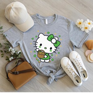 May include: A gray t-shirt featuring a cartoon character with a green bow, holding a basket of gold coins. The design includes green shamrocks and gold stars. A beige baseball cap and white sneakers are also visible.