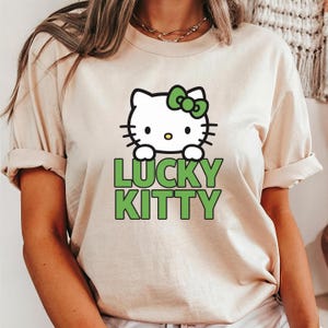 Peut inclure: T-shirt beige avec un chat de dessin anim&eacute; blanc orn&eacute; d'un n&oelig;ud vert et l'inscription "LUCKY KITTY" en vert. Le chat a des moustaches noires et un nez jaune. Le t-shirt a des manches courtes.