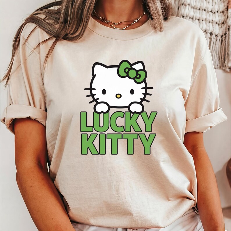 Peut inclure: T-shirt beige avec un chat de dessin anim&eacute; blanc orn&eacute; d'un n&oelig;ud vert et l'inscription "LUCKY KITTY" en vert. Le chat a des moustaches noires et un nez jaune. Le t-shirt a des manches courtes.