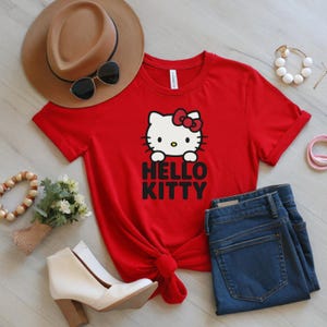 Peut inclure: Un t-shirt rouge avec un motif Hello Kitty, un n&oelig;ud rouge et le texte "HELLO KITTY" en noir. L'image comprend un chapeau marron, des lunettes de soleil, un jean bleu, des bottes blanches et des accessoires.
