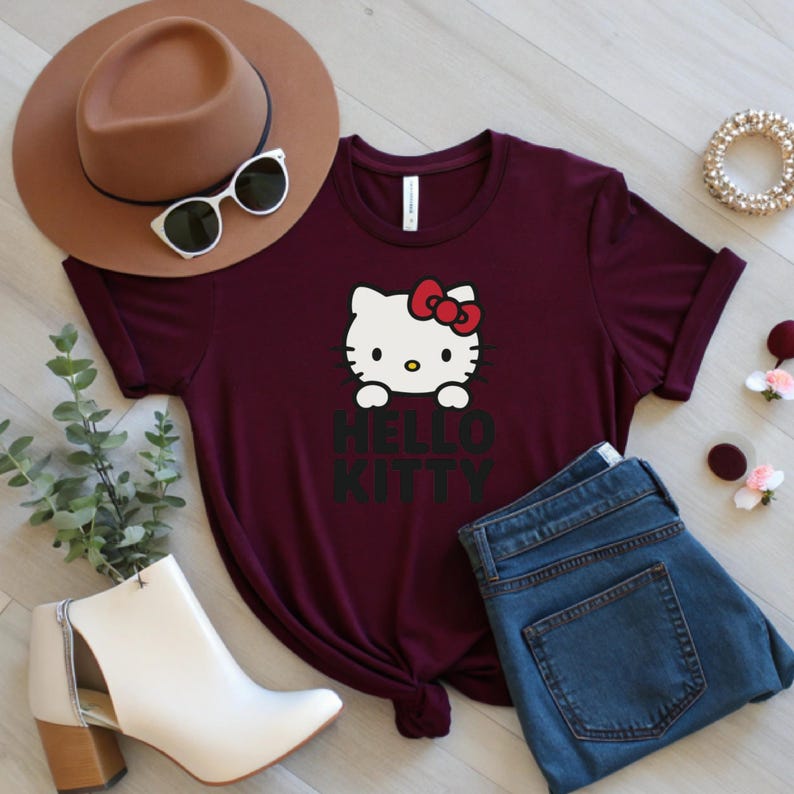 Peut inclure: T-shirt bordeaux avec un motif Hello Kitty, un n&oelig;ud rouge et l'inscription "HELLO KITTY" en noir. L'image comprend un jean bleu, un chapeau marron, des lunettes de soleil et des bottines blanches.