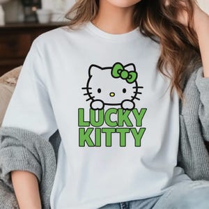Peut inclure: T-shirt blanc avec un chat de dessin anim&eacute;, un n&oelig;ud vert et l'inscription "LUCKY KITTY" en vert. Le chat a des moustaches noires et un nez jaune. Le t-shirt est de style d&eacute;contract&eacute;.
