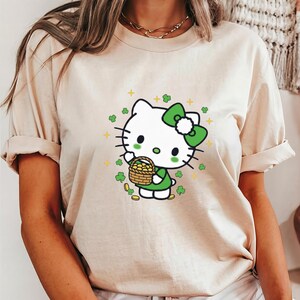 May include: A beige t-shirt featuring a cartoon character with a white head, green bow, and green dress, holding a basket of gold coins. The design includes green clovers and gold stars, creating a festive St. Patrick's Day theme.