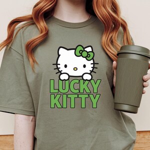 Peut inclure: T-shirt vert olive avec un chat de dessin anim&eacute; blanc, un n&oelig;ud vert et le texte "LUCKY KITTY" en vert. Un mug de voyage vert fonc&eacute; est tenu dans l'image. V&ecirc;tement de style d&eacute;contract&eacute;.