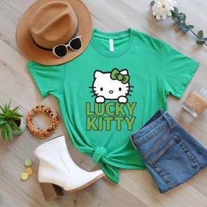Peut inclure: T-shirt vert avec un motif Hello Kitty et l'inscription "LUCKY KITTY" en vert. L'image comprend un chapeau marron, des lunettes de soleil, des bottes blanches et un jean bleu.