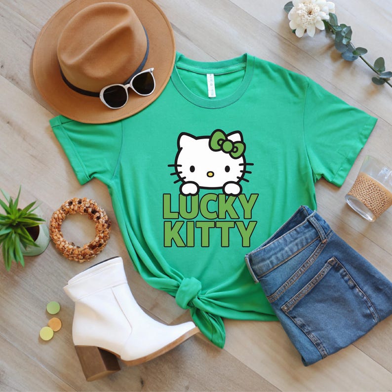 Peut inclure: T-shirt vert avec un motif Hello Kitty et l'inscription "LUCKY KITTY" en vert. L'image comprend un chapeau marron, des lunettes de soleil, des bottes blanches et un jean bleu.
