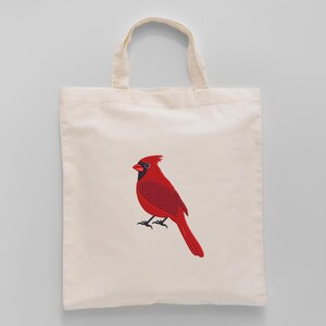 May include: A cream-colored tote bag featuring a detailed embroidered image of a vibrant red cardinal bird. The bird has black accents and is perched on a branch. The bag has two handles.