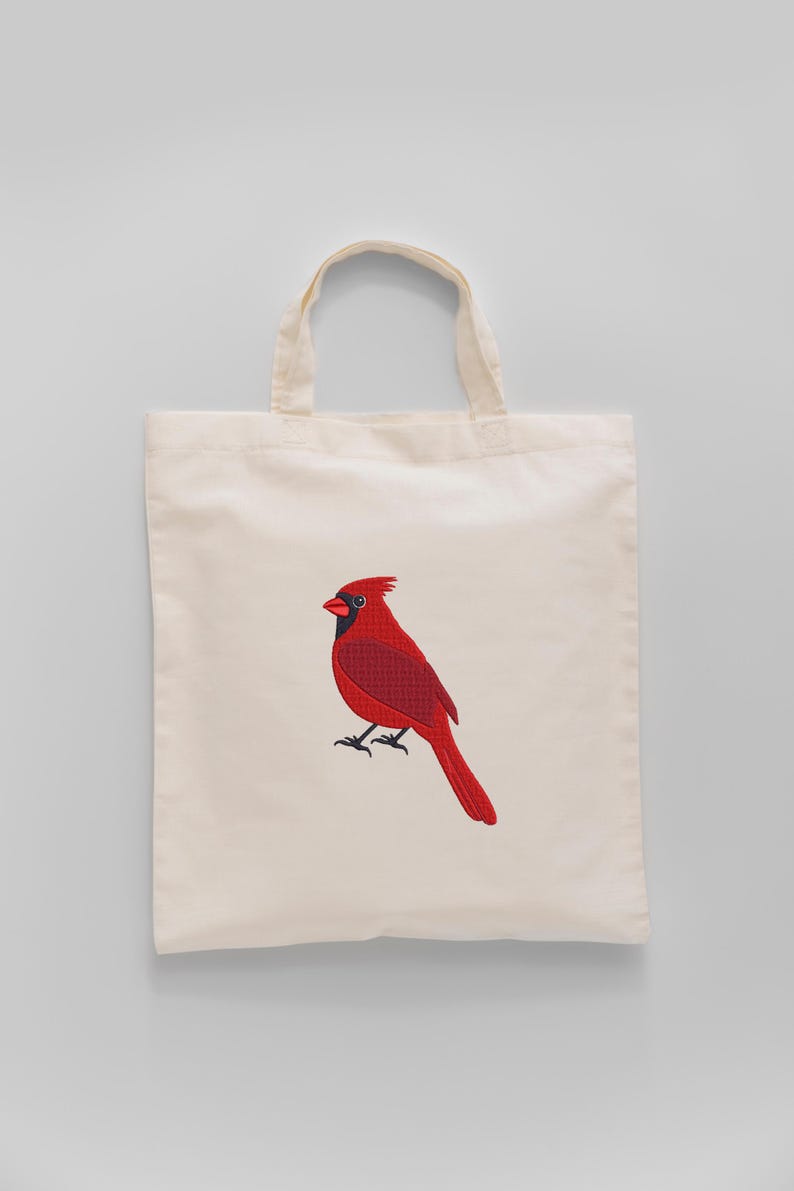 May include: A cream-colored tote bag featuring a detailed embroidered image of a vibrant red cardinal bird. The bird has black accents and is perched on a branch. The bag has two handles.