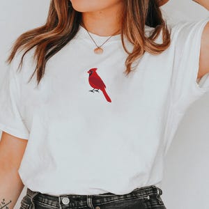 May include: White t-shirt featuring a red cardinal bird design. The bird is embroidered on the front of the shirt. The shirt has short sleeves and a crew neck. The overall style is casual and comfortable.