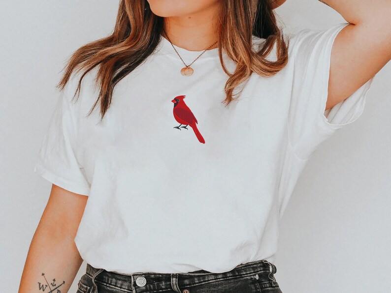 May include: White t-shirt featuring a red cardinal bird design. The bird is embroidered on the front of the shirt. The shirt has short sleeves and a crew neck. The overall style is casual and comfortable.