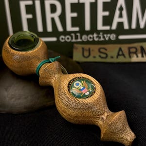 May include: A wooden pipe with a green glass bowl and a decorative wooden handle. The handle features a U.S. Army emblem. The background includes the text "FIRETEAM collective" and a U.S. Army patch.