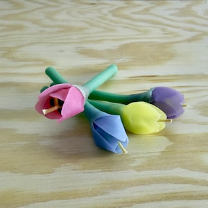 May include: Four tulip-shaped candles in pink, blue, yellow, and purple. Each candle has a green stem and a small, wooden stamen. The candles are arranged on a light-coloured wooden surface.